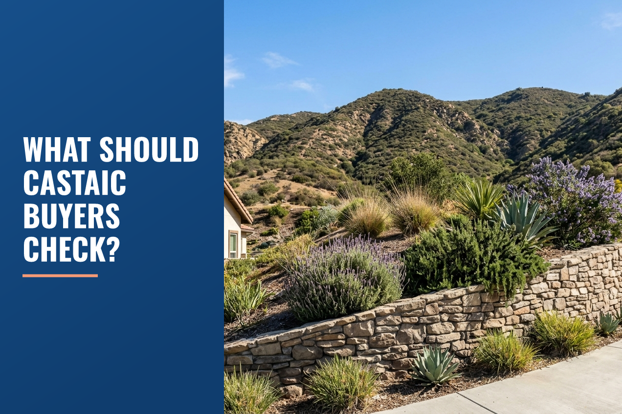 A street-level view of a stone retaining wall, drought-tolerant landscaping, and foothills under a clear sky in a suburban setting.